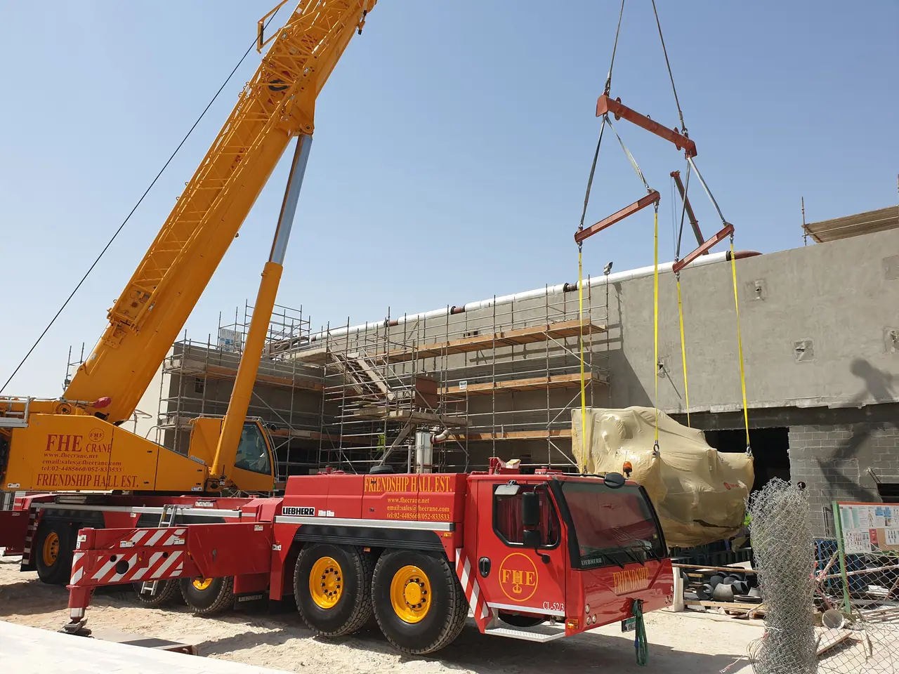 All-terrain crane in Friendship Hall livery with wrapped plant equipment on a spreader, beside a building