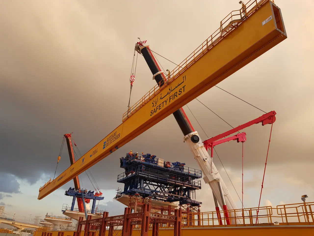A mobile crane boom crossing a yellow bridge-launching girder marked SAFETY FIRST, under a dusk sky