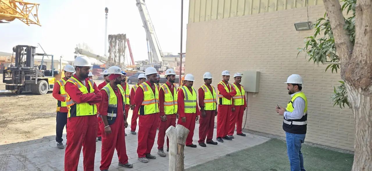 A briefing beside a building wall, with a forklift and a mobile crane in the yard behind the crew