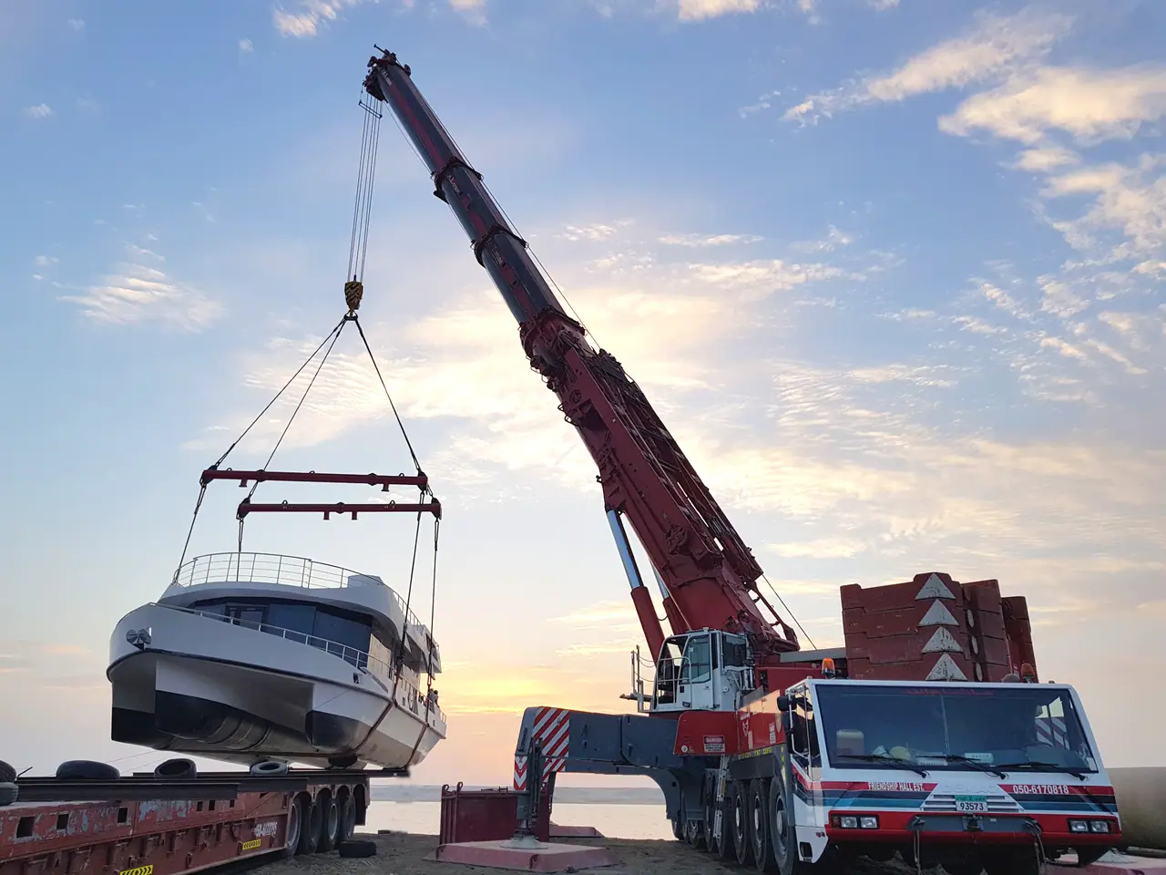 Red mobile crane with a yacht on the hook above a lowbed trailer, at sunset