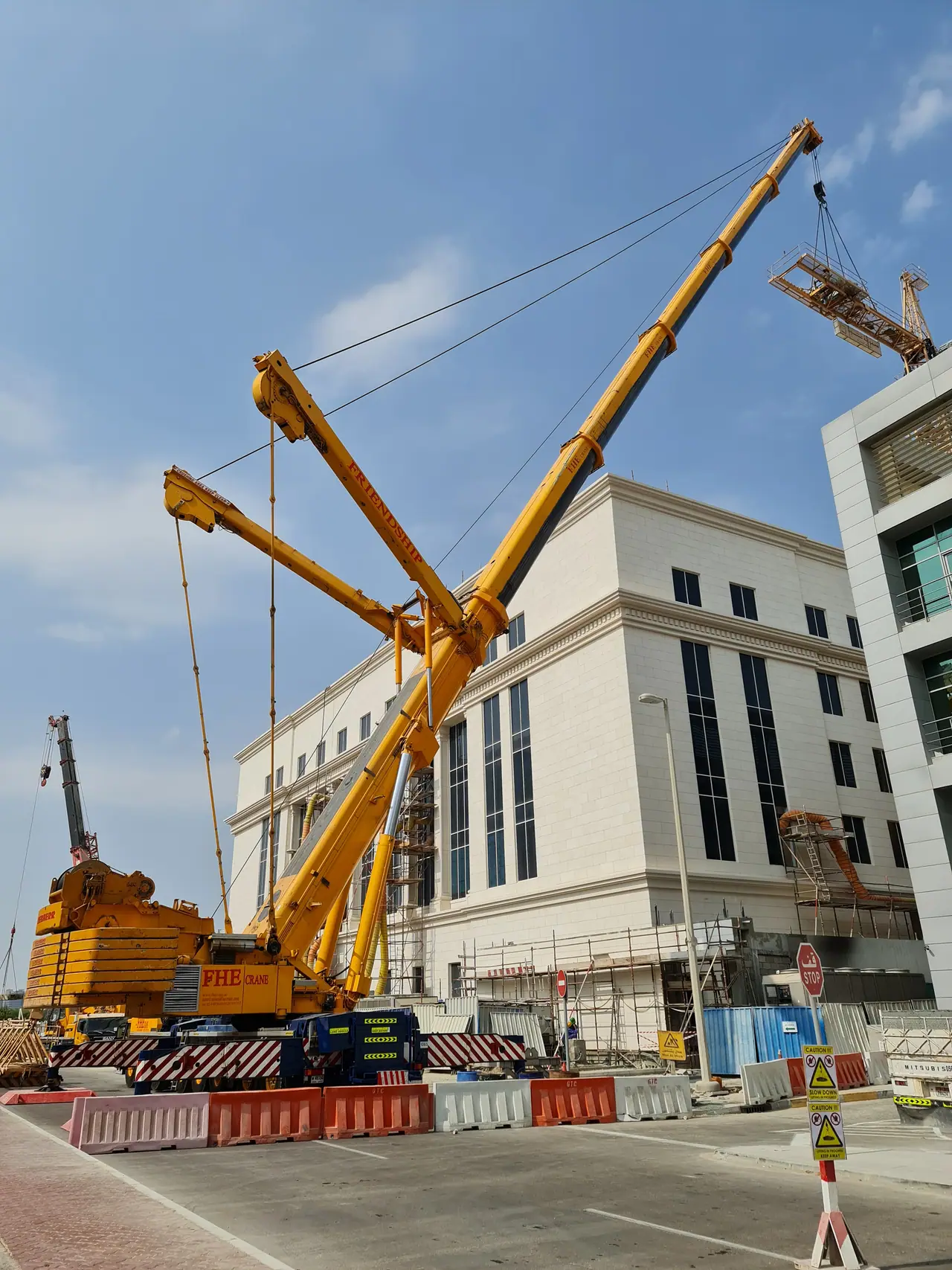 Yellow FHE crane on a luffing jib lifting a steel lattice section beside a stone-clad building