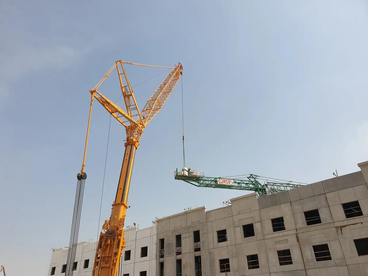 Yellow luffing-jib crane lifting a green tower-crane jib section beside a concrete frame