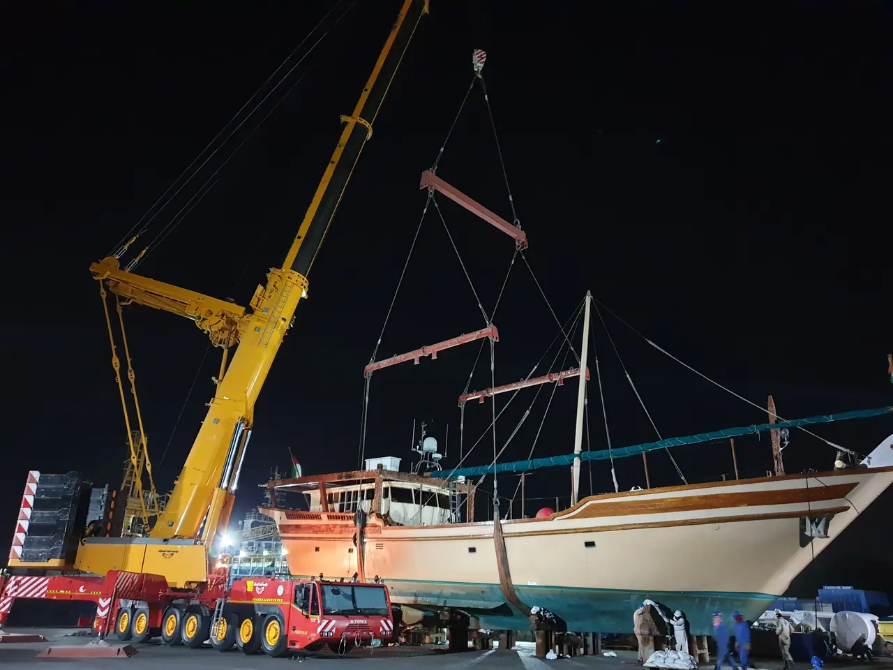 Yellow crane with a wooden dhow rigged to spreader bars, at night under floodlights