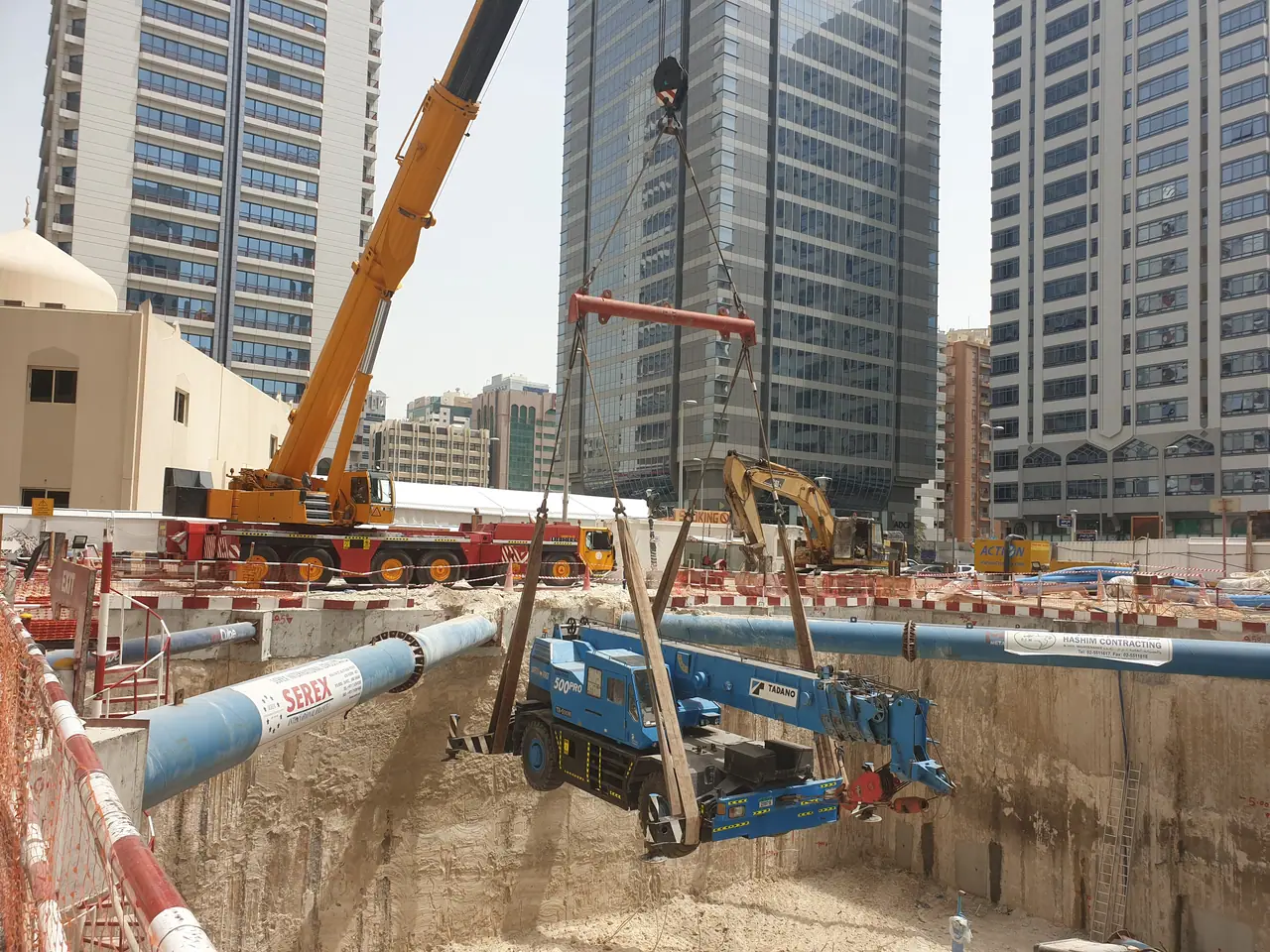 Mobile crane lifting a smaller blue crane out of a deep excavation between city towers