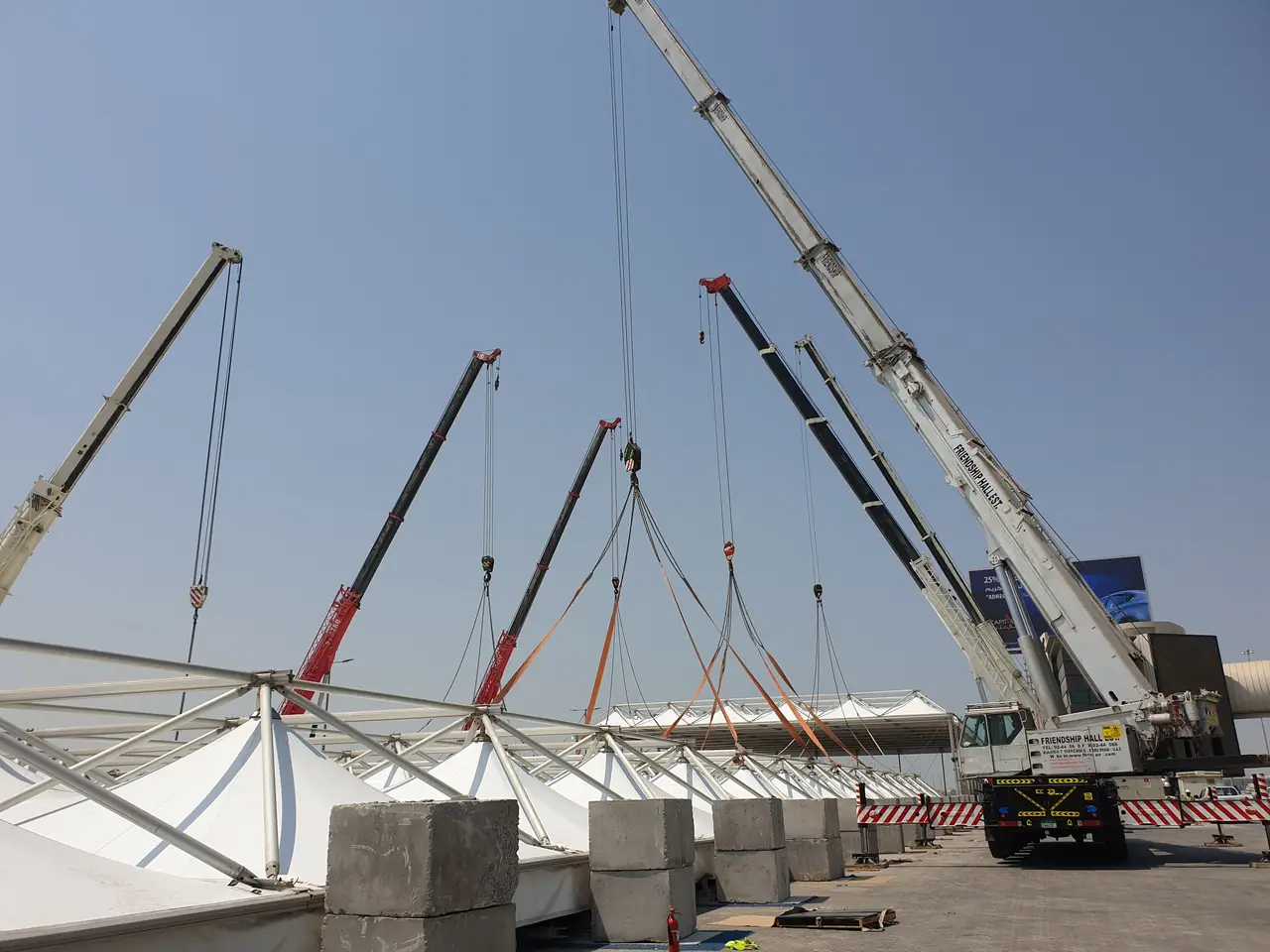 Several mobile cranes rigged to a tensile fabric roof, with a white Friendship Hall crane in front